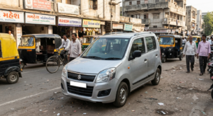 A silver Maruti Suzuki WagonR parked on a busy street in India, surrounded by auto-rickshaws, pedestrians, and shops with Gujarati and English signage.